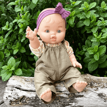 Baby doll with a purple headband and khaki overalls sitting on a log with green plants in the background