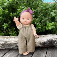 Doll wearing a purple headband and khaki overalls standing on a wooden surface with greenery in the background