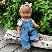 Doll wearing blue overalls with a peach headband sitting on a log with greenery in the background