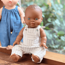 Doll wearing a beige overalls sitting on a wooden surface with another doll in the background.