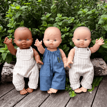 Three dolls wearing overalls standing on a wooden surface with green foliage in the background